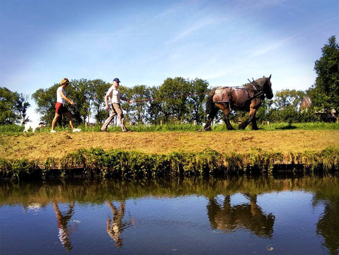 Foto van een werkpaard met twee begeleiders op een jaagpad langs de Hollandse IJssel, gezien vanaf een trekschuit.