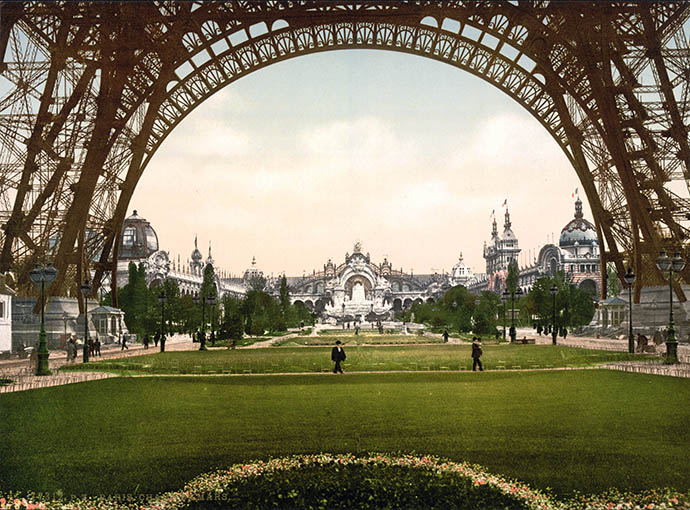 Een doorkijkje richting de entree van de Parijse wereldtentoonstelling, door de bogen van de Eiffeltoren, gebouwd ter gelegenheid van de wereldtentoonstelling van 1889. Fotochroom uit 1889 [Publiek domein].